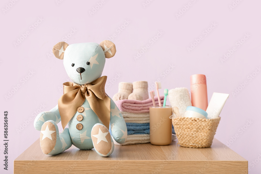 Toy bear with baby clothes and accessories on table near lilac wall