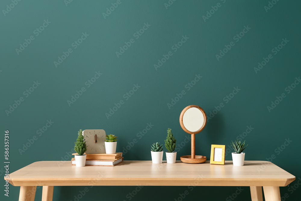 Artificial plants with books, clock and mirror on table near green wall