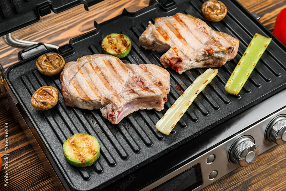 Modern electric grill with steaks and vegetables on wooden background, closeup