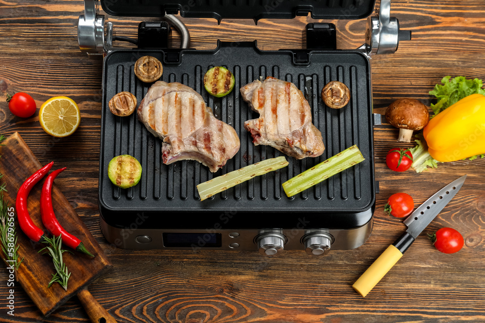 Modern electric grill with steaks and vegetables on wooden background