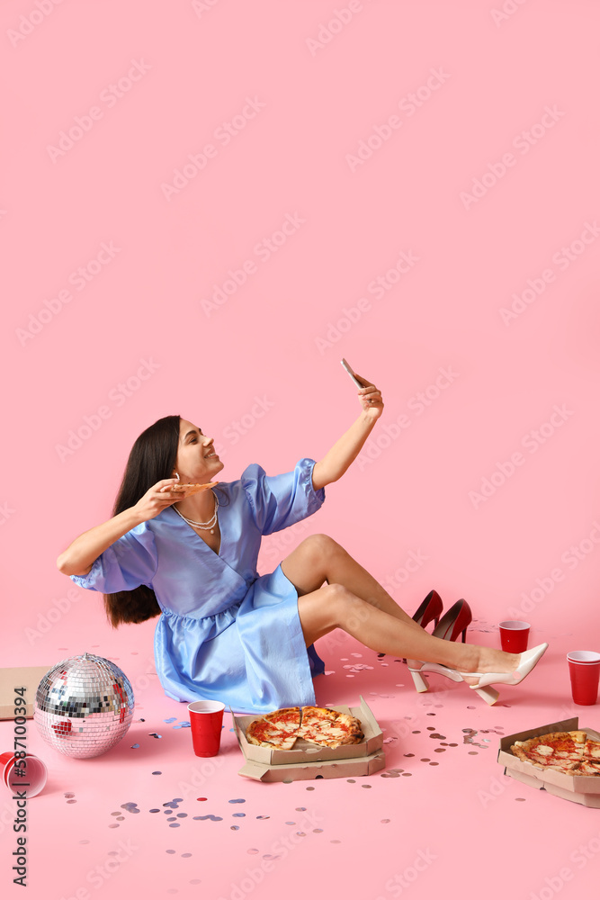 Young woman with tasty pizza, cups and disco ball taking selfie on pink background