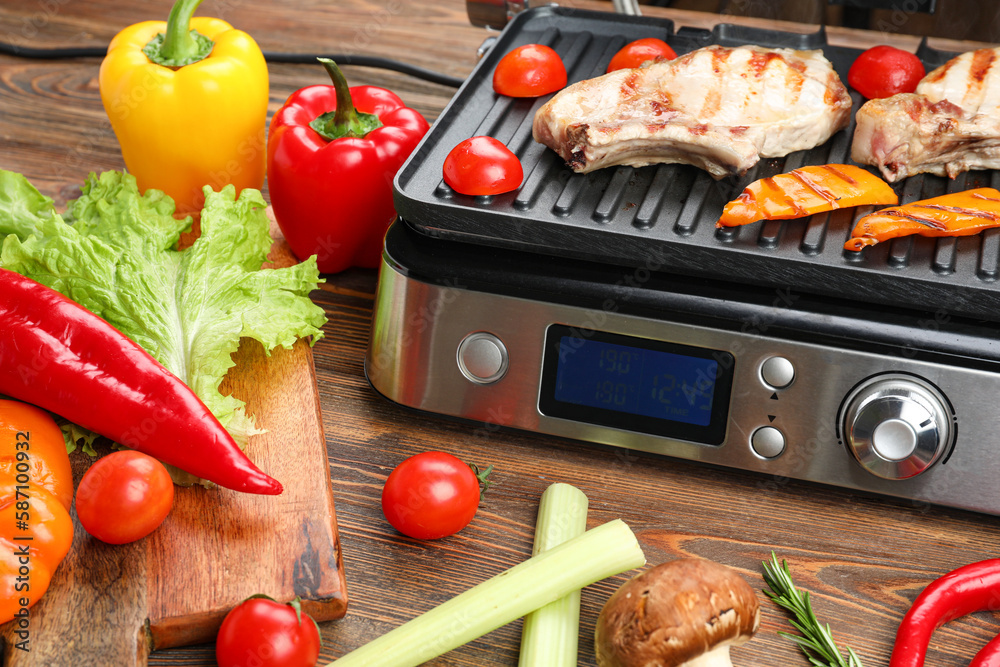 Modern electric grill with steaks and vegetables on wooden background