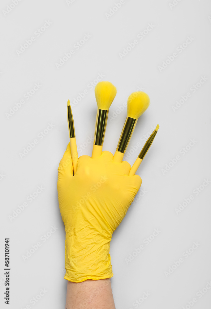 Beautician in rubber glove with makeup brushes on light background