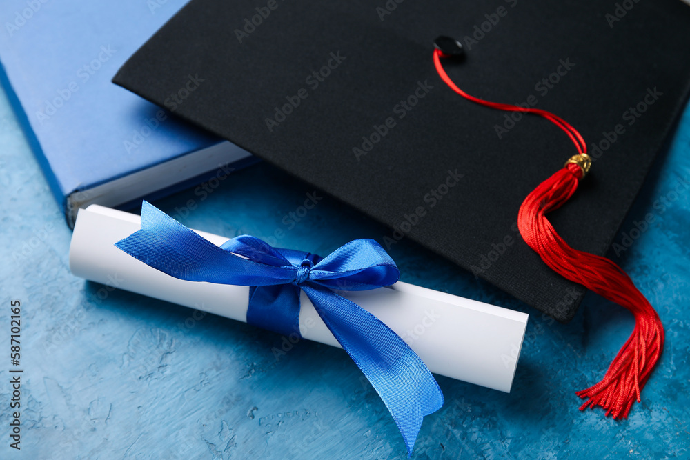 Diploma with ribbon, graduation hat and book on blue table