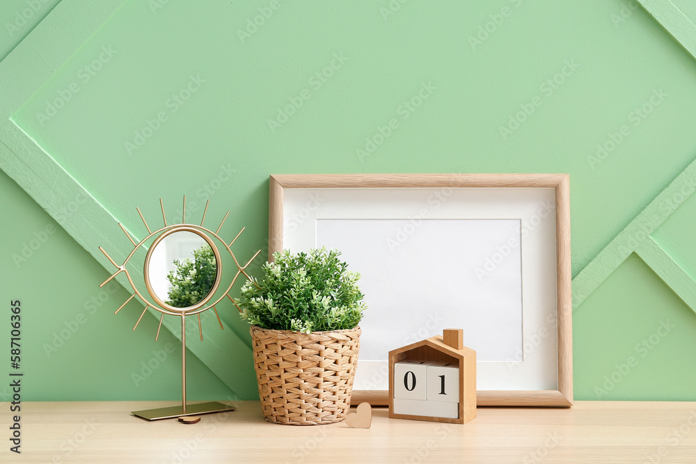 Artificial houseplant with mirror, calendar and blank frame on table near green wall