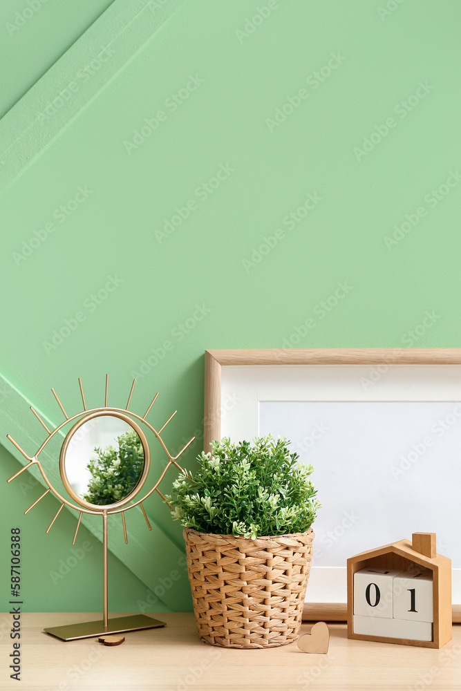 Artificial houseplant with mirror, calendar and blank frame on table near green wall