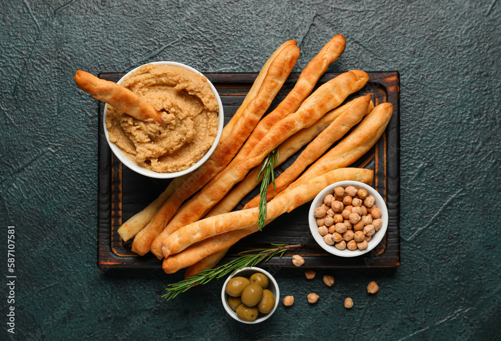 Wooden board with tasty Italian Grissini, hummus and chickpeas on dark background