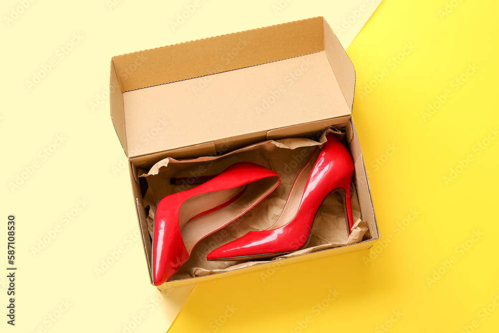 Cardboard box with high heeled shoes on color background