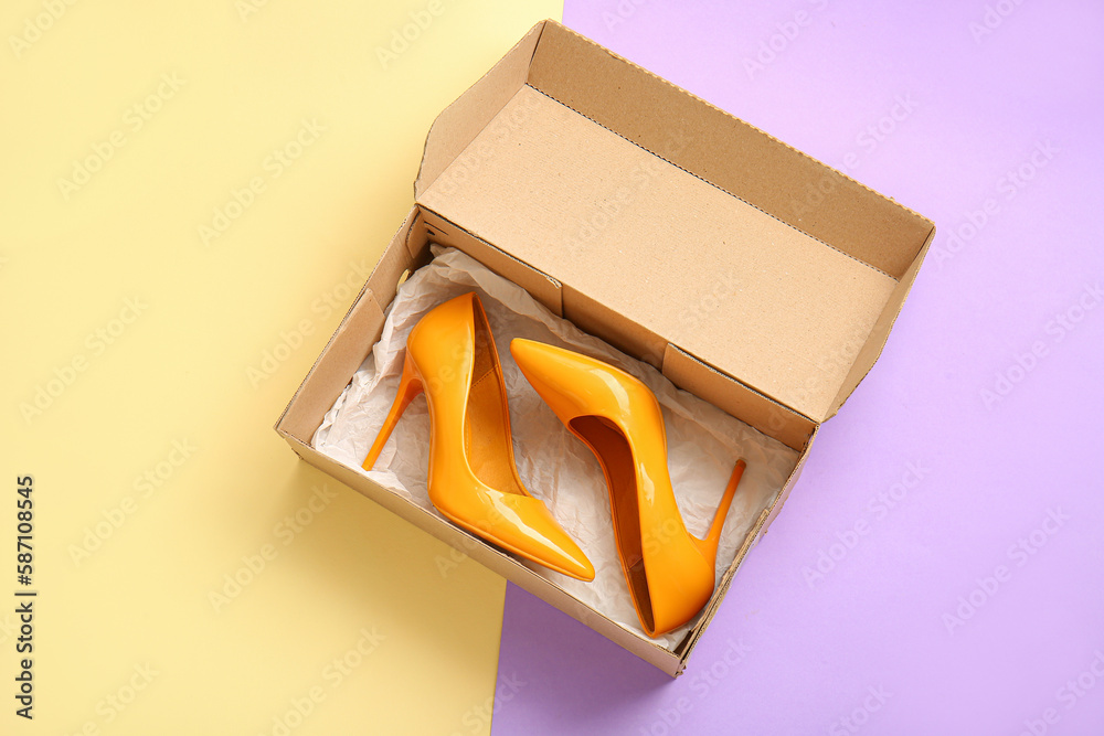 Cardboard box with high heeled shoes on color background