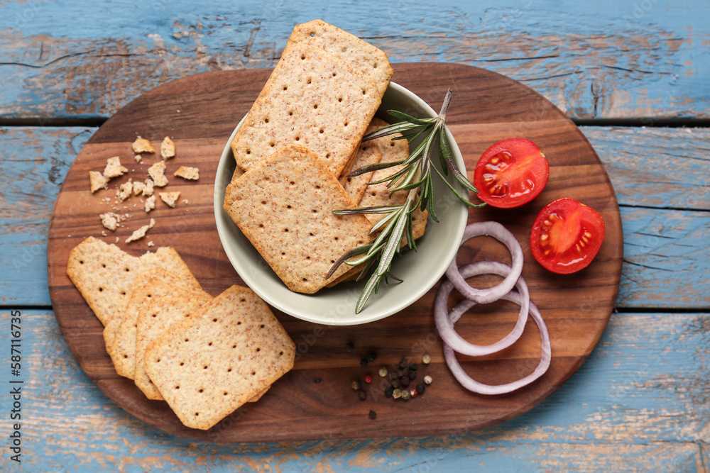 Board with tasty crackers, rosemary, onion and tomatoes on blue wooden background