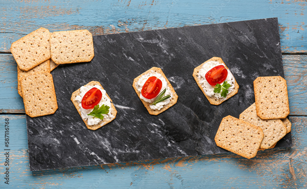 Slate board with tasty crackers, cream cheese and tomatoes on blue wooden background