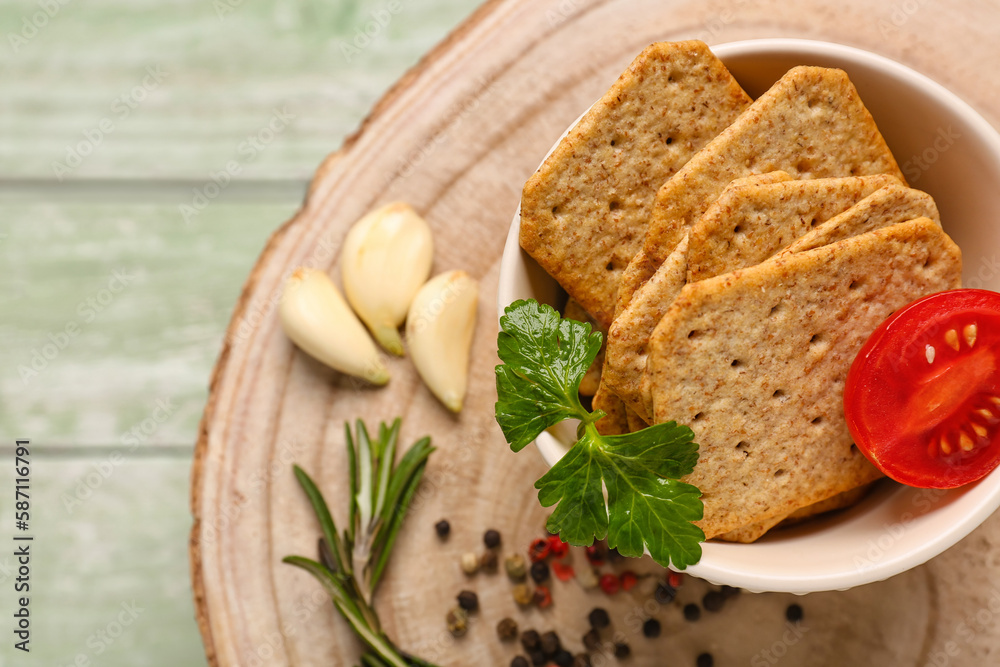 Stand with bowl of tasty crackers, garlic, peppercorn and tomato on green wooden background