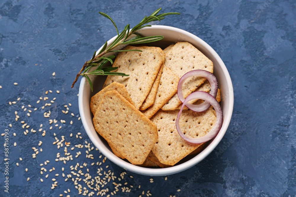 Bowl of tasty crackers with onion rings, rosemary and sesame seeds on blue background