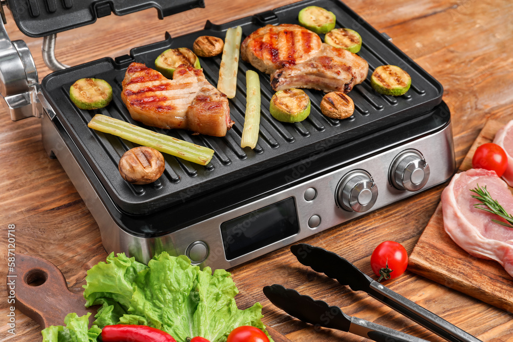 Modern electric grill with tasty steaks and vegetables on wooden background