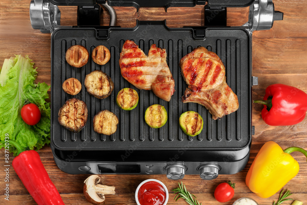 Modern electric grill with tasty steaks and vegetables on wooden background