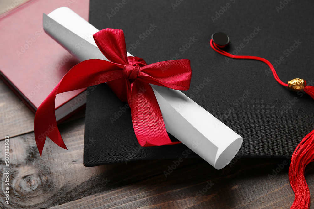 Diploma with red ribbon, graduation hat and book on wooden table