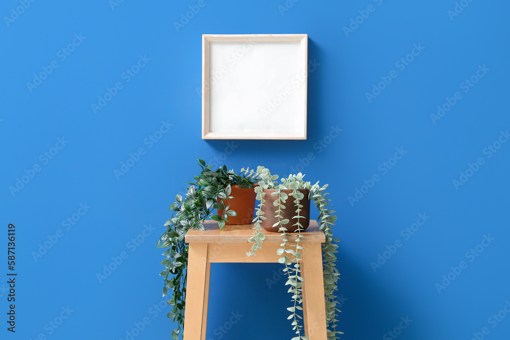 Table with artificial houseplants and blank frame on blue wall