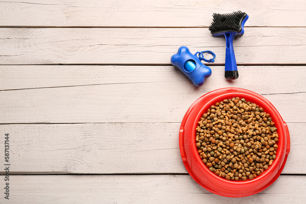 Bowl of dry pet food, waste bags and grooming brush on white wooden background