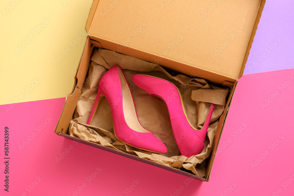 Cardboard box with high heeled shoes on color background