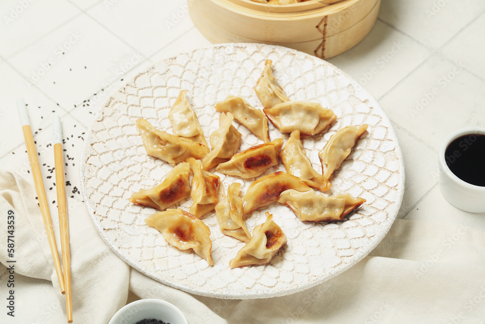 Plate with tasty Chinese jiaozi, sesame and bamboo steamer on white tile background