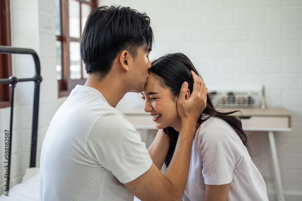 Asian husband kissing his wife while sitting on bed in bedroom at home. 