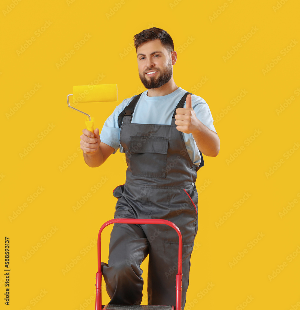 Male worker with paint roller and ladder showing thumb-up on yellow background