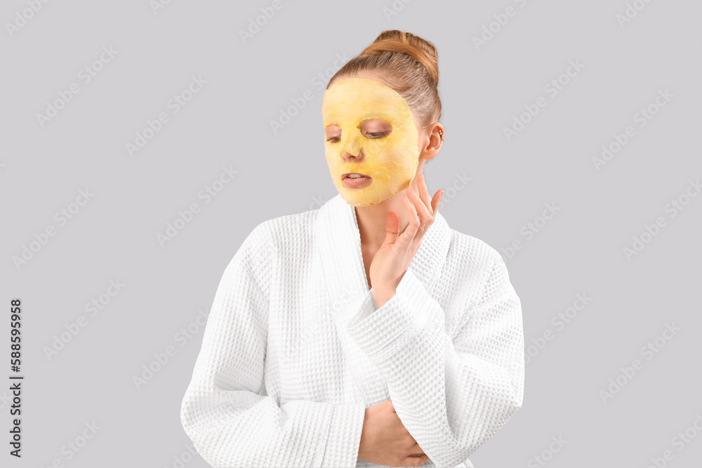 Young woman in bathrobe with turmeric sheet mask on grey background