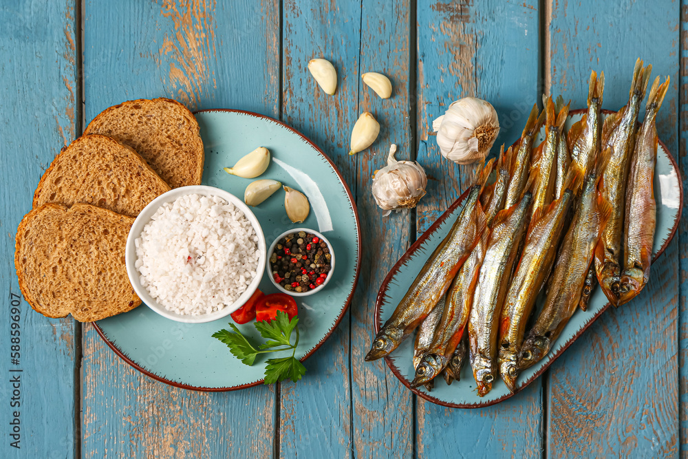 Plates with tasty smoked capelin, bread pieces, garlic and sea salt on blue wooden background