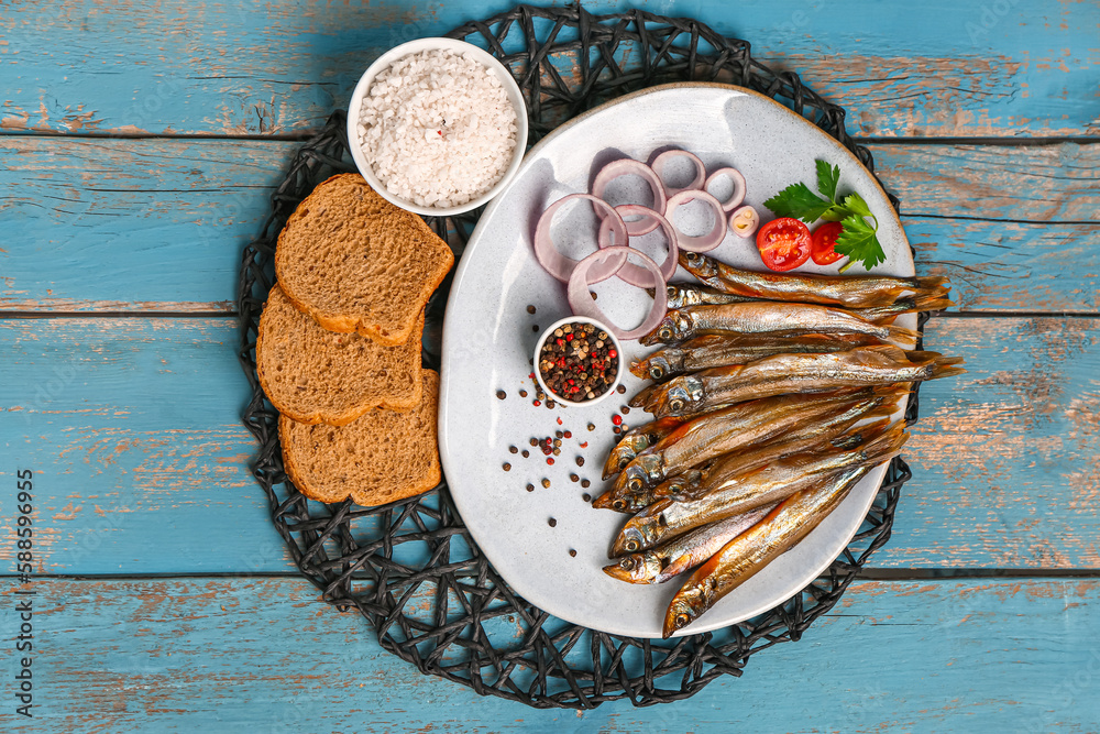 Plate with tasty smoked capelin and bread pieces on blue wooden background