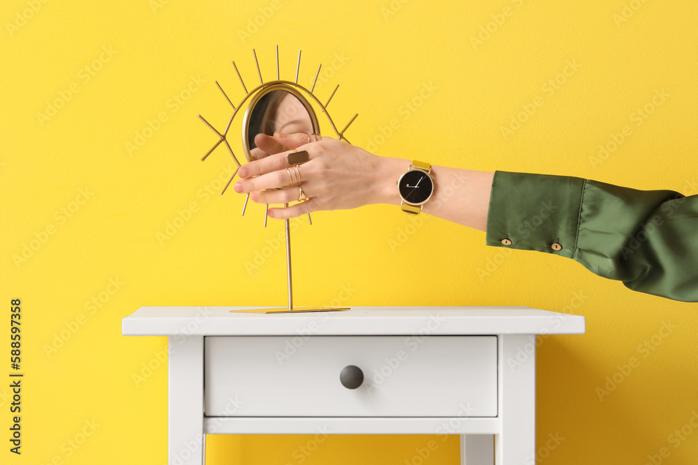 Woman with golden wristwatch and mirror on table near yellow wall