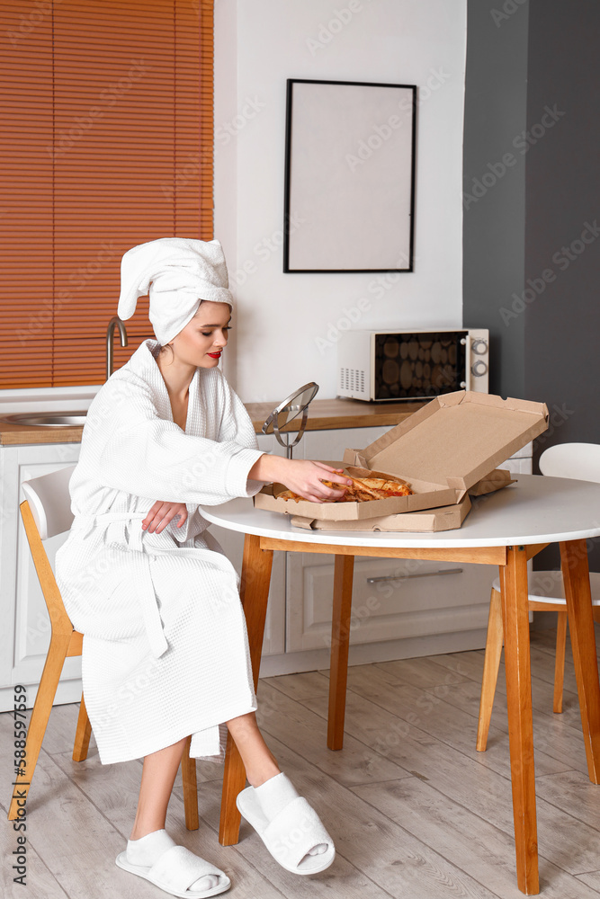 Young woman in bathrobe with tasty pizza and mirror on table at home