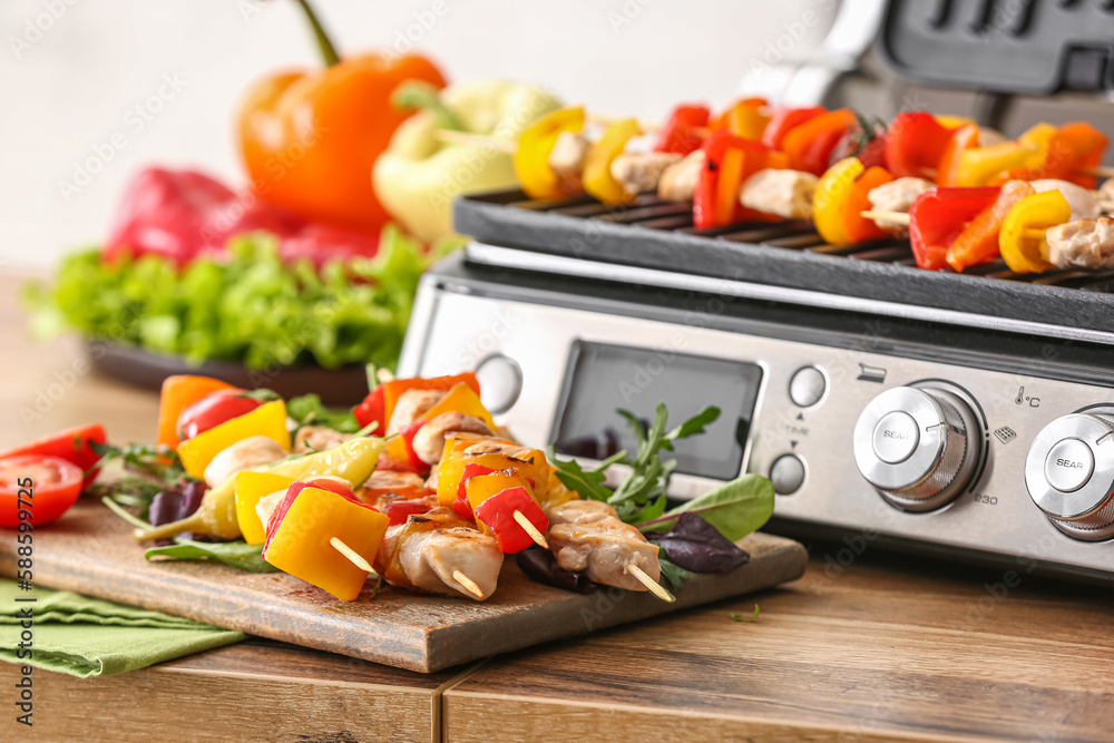 Wooden board with tasty chicken skewers, vegetables and modern electric grill on table in kitchen