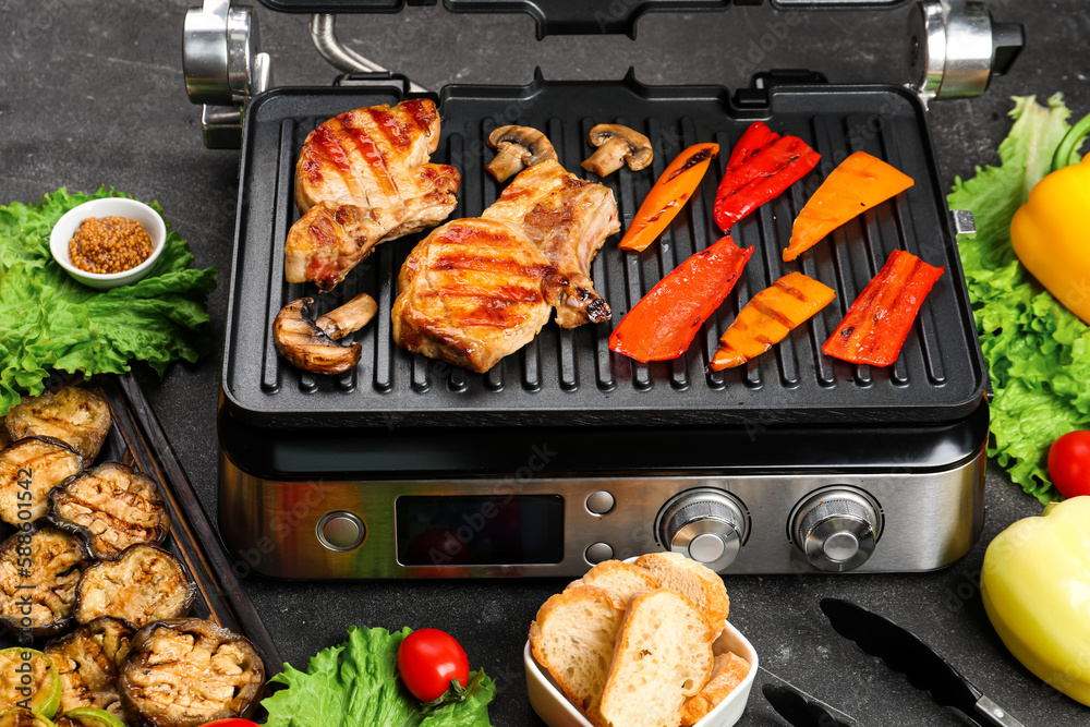 Modern electric grill with tasty steaks and vegetables on dark background