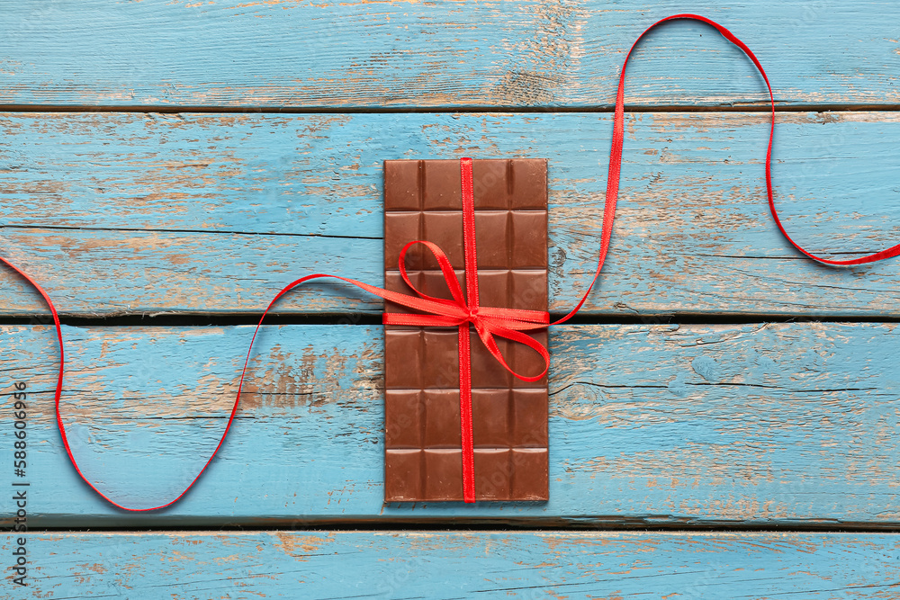 Tasty chocolate bar tied with red ribbon on color wooden background