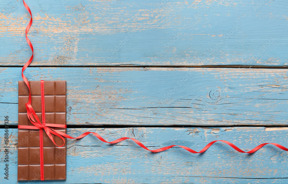 Tasty chocolate bar tied with red ribbon on color wooden background