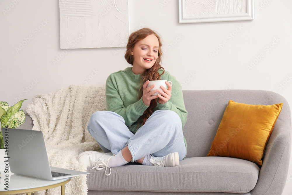 Beautiful redhead woman with cup of tea sitting on sofa at home