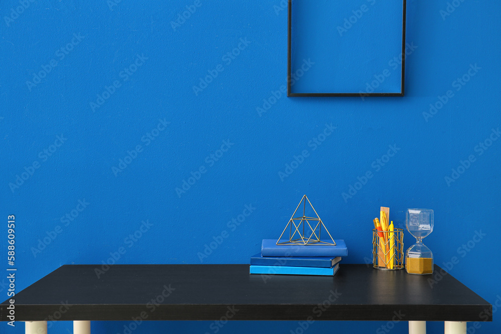 Workplace with stationery, books and hourglass near blue wall