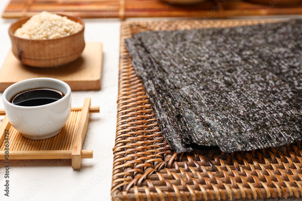 Wicker mat with nori sheets and soy sauce on light table, closeup