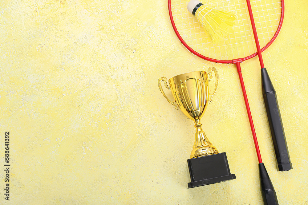 Gold cup with badminton rackets and shuttlecock on yellow background