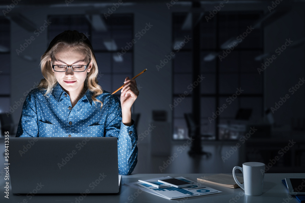 Attractive blonde working on laptop in dark office. Mixed media