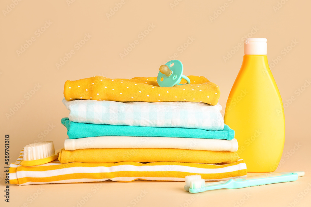 Stack of baby clothes, accessories and bottle of cosmetic product on color background