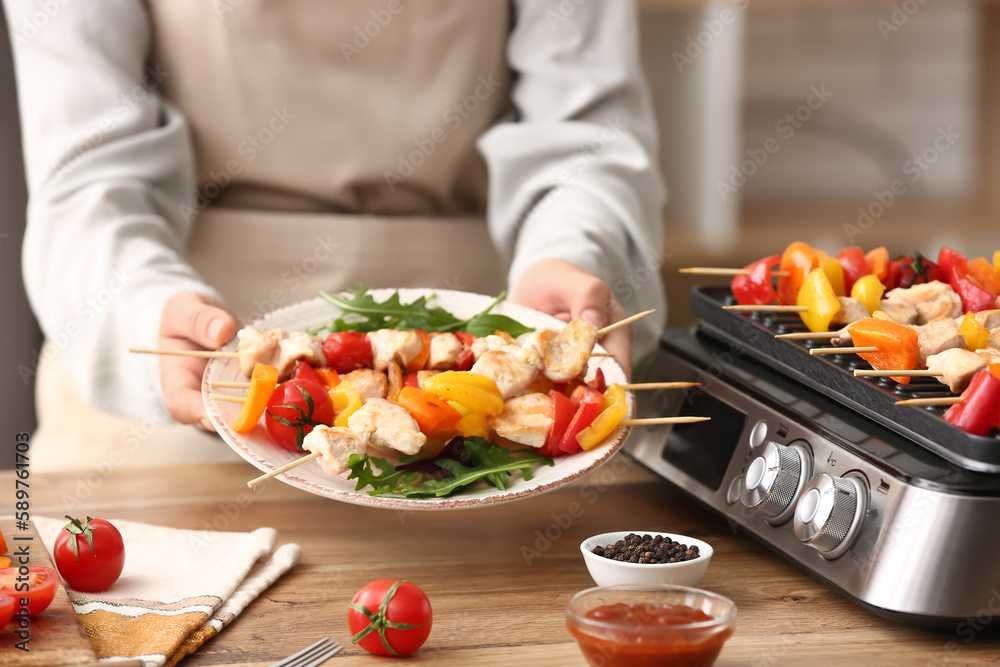 Woman holding plate with grilled chicken skewers and vegetables in kitchen, closeup