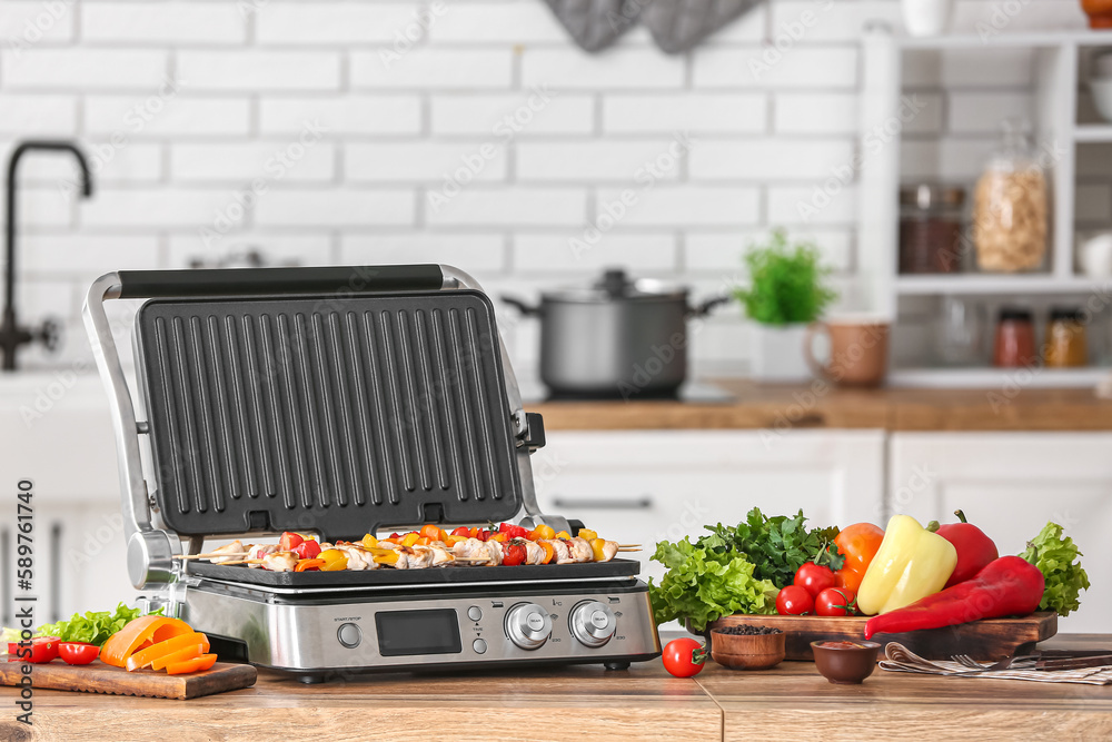 Modern electric grill with tasty chicken skewers and vegetables on table in kitchen