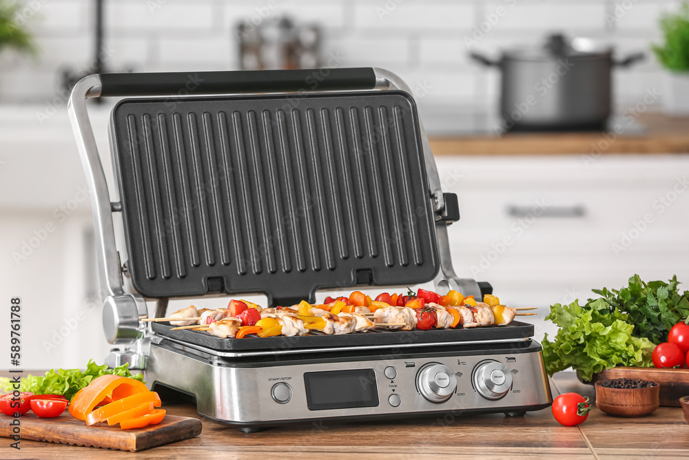 Modern electric grill with tasty chicken skewers and vegetables on table in kitchen