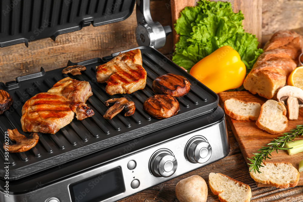 Modern electric grill with tasty steaks and vegetables on wooden background, closeup