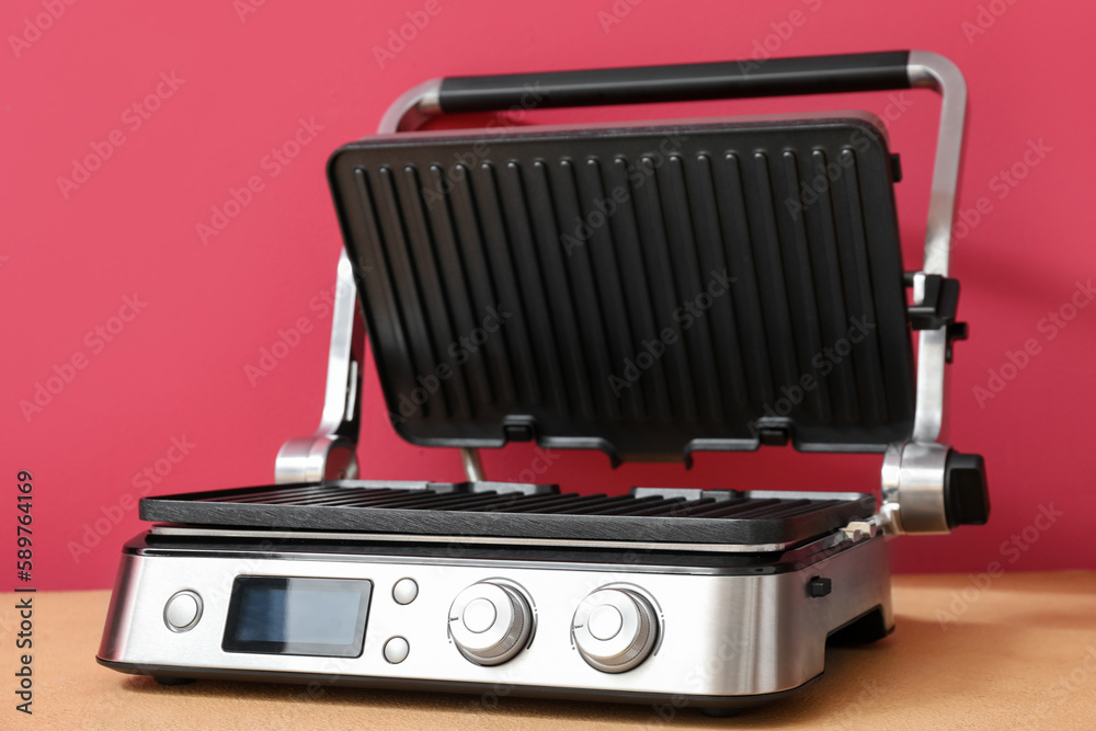 Modern electric grill with open lid on beige table against red background