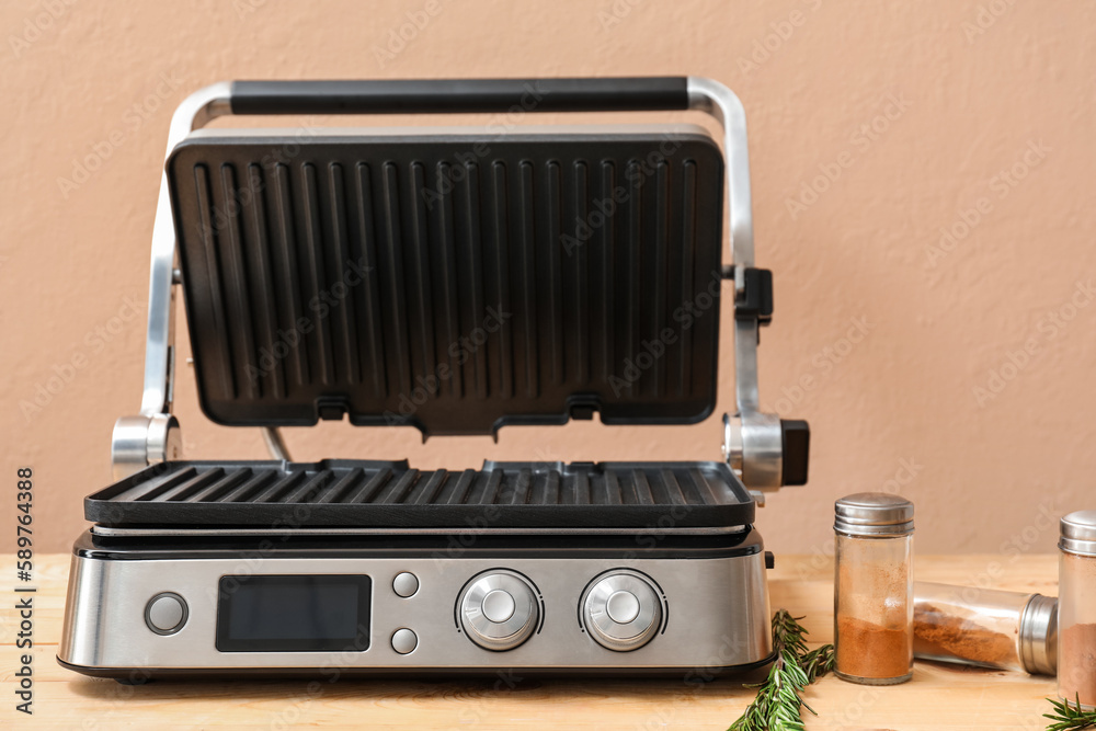 Modern electric grill with open lid, spices and rosemary on wooden table against beige background