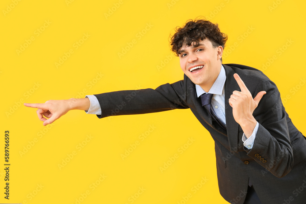 Laughing young businessman showing loser gesture and pointing at something on yellow background