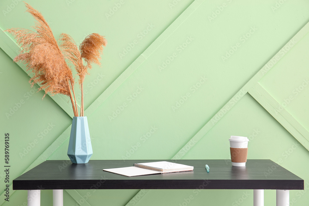 Workplace with notebook, pampas grass in vase and cup of coffee near green wall