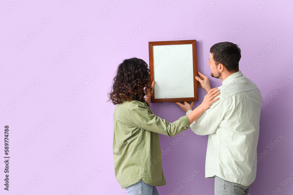 Young couple hanging blank frame on lilac wall